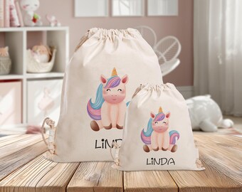 Könnte beinhalten: Zwei beigefarbene Kordelzugbeutel mit einem Cartoon-Einhorn-Design. Die Einhörner haben rosa Körper, braune Hufe und bunte Mähnen und Schwänze. Der Name "LINDA" ist in Schwarz auf jeder Tasche aufgedruckt. Eine Tasche ist größer als die andere.