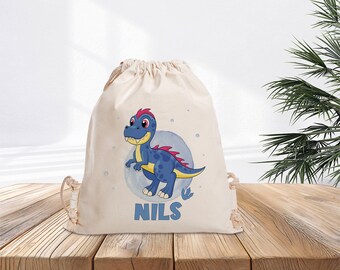 Könnte beinhalten: Eine weiße Kordeltasche mit einer blauen Dinosaurier-Illustration und dem Namen "NILS" auf der Vorderseite.