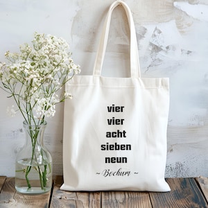 Peut inclure: Sac fourre-tout crème avec texte noir "vier vier acht sieben neun ~ Bochum ~". Le sac a deux anses et est fait d'un tissu robuste. Un vase en verre transparent avec des fleurs blanches est à gauche.