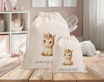 Könnte beinhalten: Zwei beige Kordelzugbeutel mit einem Einhorn-Design. Die Taschen zeigen ein sitzendes Einhorn mit einem bunten Horn und dem Namen "ANN-KATHRIN" in Schwarz. Die Taschen befinden sich auf einer Holzoberfläche.