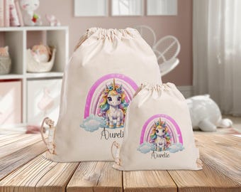 Könnte beinhalten: Zwei weiße Kordelbeutel mit einem Einhorn- und Regenbogen-Design. Der größere Beutel hat den Namen "Aurelie" aufgedruckt. Der kleinere Beutel hat das gleiche Design, aber einen anderen Namen.