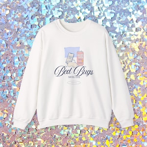 May include: White crewneck sweatshirt with the text "Bed Bugs SOCIAL CLUB" in a script font. The design includes illustrations of a phone, a can, and a pillow. The sweatshirt is a casual, comfortable style.