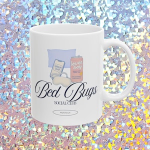 May include: White ceramic mug featuring the text "Bed Bugs Social Club" in a script font, with illustrations of a phone, eye mask, pillow, and a can of "Lover Boy" strawberry lemonade. The word "Montauk" is also printed on the mug.