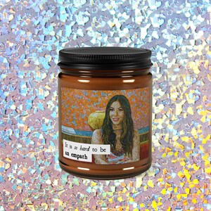 May include: A brown candle jar with a black lid. The label features a mosaic-style portrait of a woman and the text "It is so hard to be an empath."
