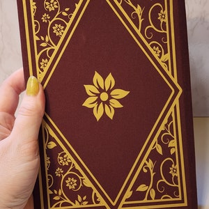 以下が含まれることがあります： 金色の花の模様と金色のひし形の枠が付いた、えんじ色のハードカバーのジャーナル。ジャーナルには、ひし形の真ん中に金色の花が描かれている。