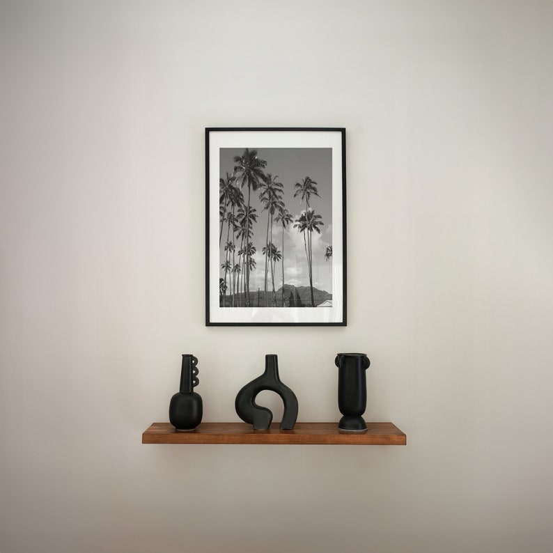 May include: A black and white photograph of palm trees in a tropical setting is framed in a black frame and displayed on a wall above a wooden shelf. Three black ceramic vases are arranged on the shelf.