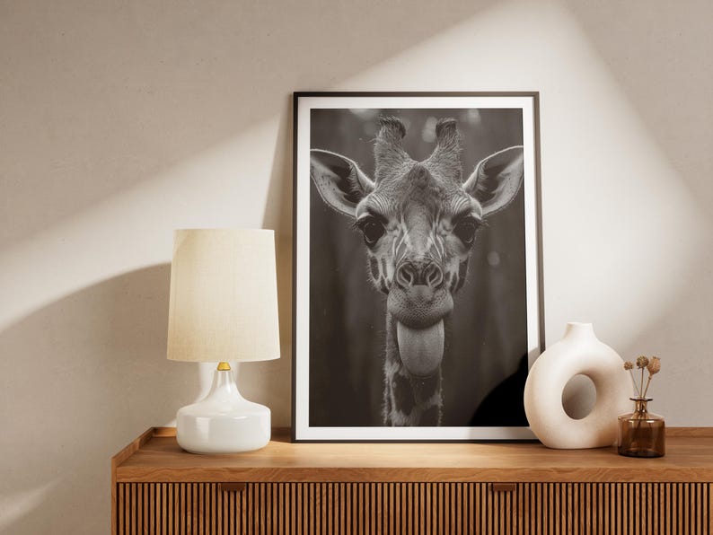 May include: Black and white photograph of a giraffe's head and neck. The giraffe is looking directly at the camera with its tongue sticking out. The photograph is framed and hanging on a wall above a wooden cabinet.