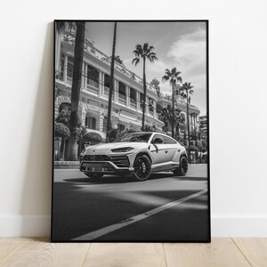 May include: A black and white photograph of a white SUV driving down a street lined with palm trees and a building in the background.