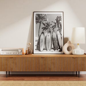 May include: A wooden media console with a black and white photo of surfboards and palm trees hanging above it. The console has a stereo receiver on top and a white lamp with a white shade on the right side.