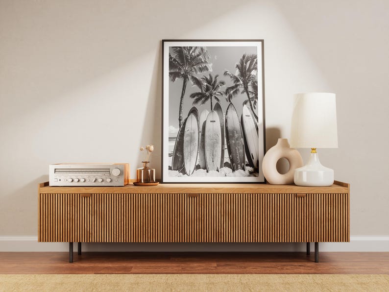 May include: A wooden media console with a black and white photo of surfboards and palm trees hanging above it. The console has a stereo receiver on top and a white lamp with a white shade on the right side.