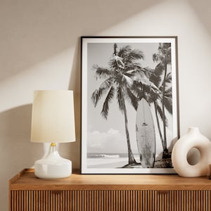 May include: Black and white photograph of palm trees and a surfboard on a beach. The photo is framed and displayed on a wooden shelf with a white lamp and a white vase.