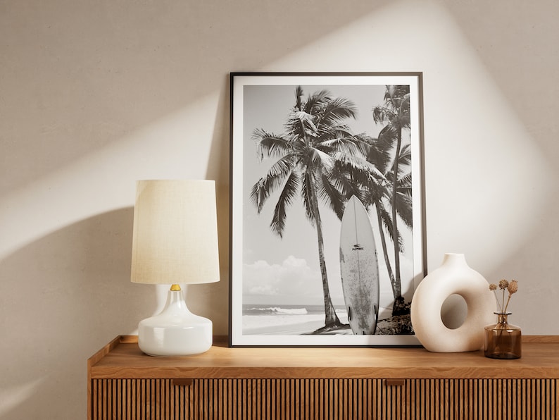 May include: Black and white photograph of palm trees and a surfboard on a beach. The photo is framed and displayed on a wooden shelf with a white lamp and a white vase.