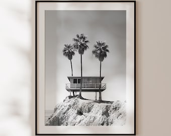 Puede incluir: Fotografía en blanco y negro de una pequeña casa de madera sobre pilotes situada en un acantilado con vistas al océano. Tres palmeras se alzan detrás de la casa.