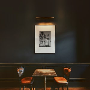 May include: A black and white photograph of palm trees in a frame hangs on a dark wall above a table with two brown leather chairs. The table has a dark wood top and a gold metal base.