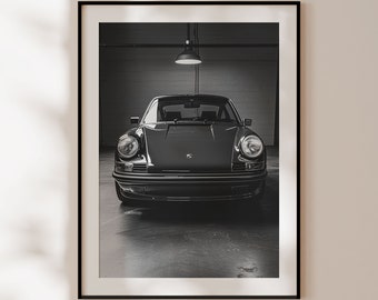 May include: A black and white photograph of a classic Porsche 911 parked in a garage. The car is facing the camera and the headlights are on.