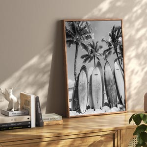 May include: Black and white photograph of surfboards leaning against palm trees on a beach. The surfboards are lined up in a row and have a vintage look.