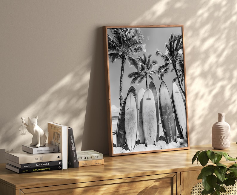 May include: Black and white photograph of surfboards leaning against palm trees on a beach. The surfboards are lined up in a row and have a vintage look.