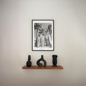 May include: Black and white photograph of surfboards leaning against palm trees on a beach. The photo is framed in a black frame and is displayed on a shelf with three black ceramic vases.
