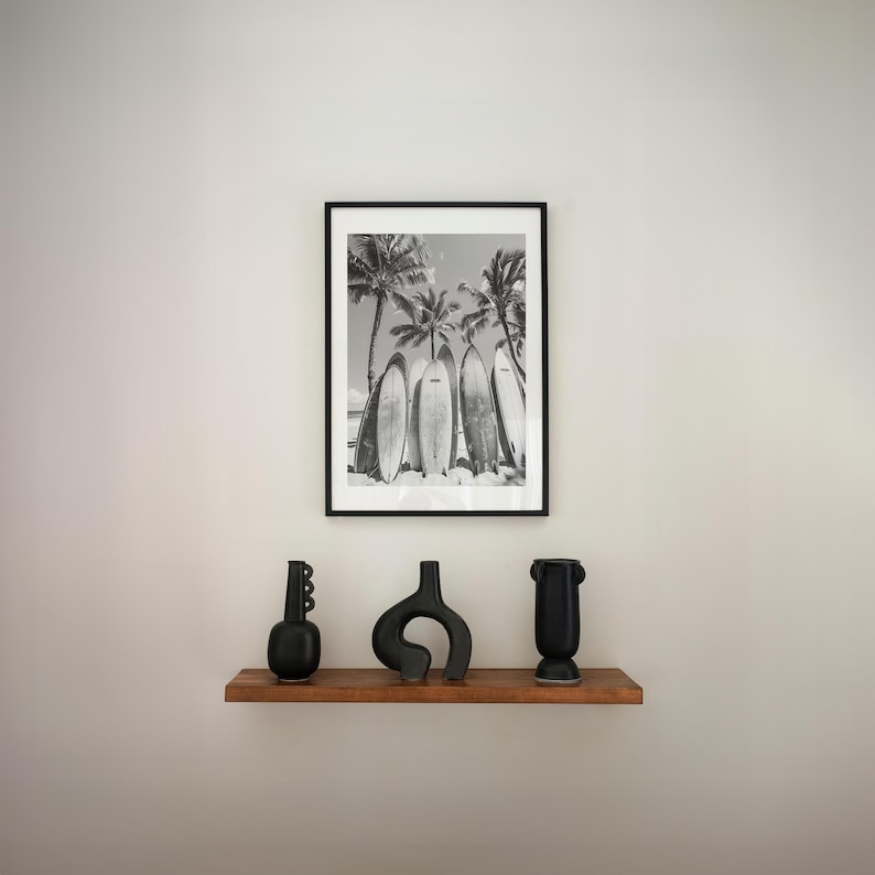 May include: Black and white photograph of surfboards leaning against palm trees on a beach. The photo is framed in a black frame and is displayed on a shelf with three black ceramic vases.