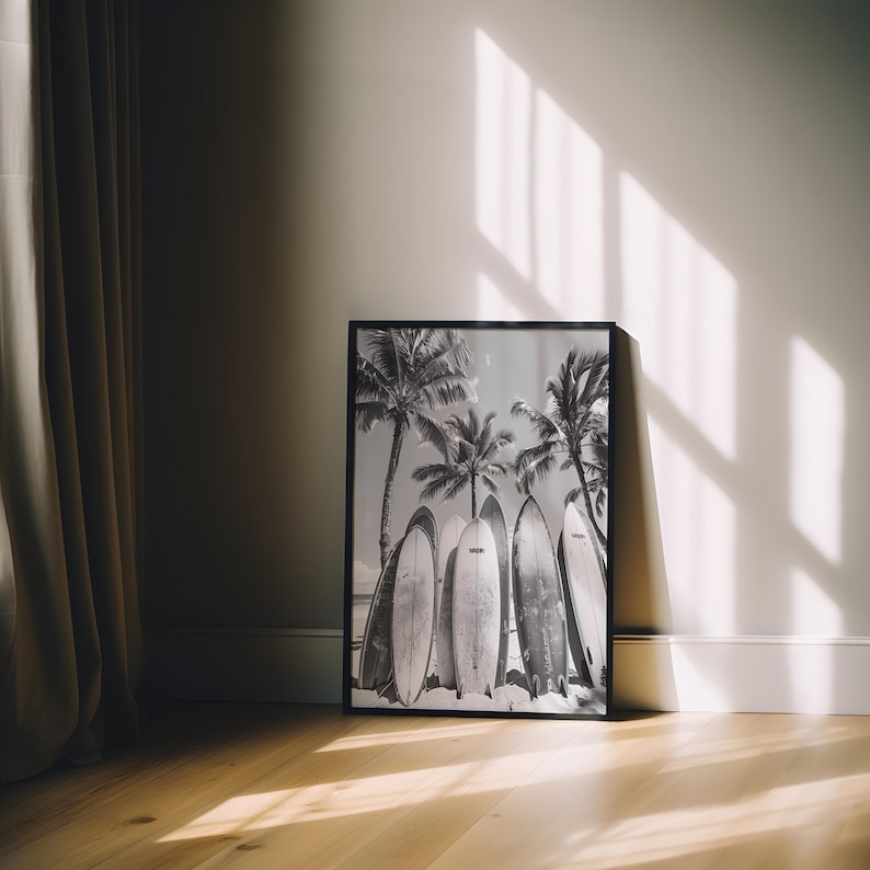 May include: Black and white photograph of surfboards leaning against palm trees on a beach. The photo is framed in a black frame.