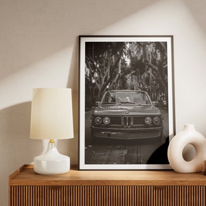 Puede incluir: Fotograf&iacute;a en blanco y negro de un coche cl&aacute;sico conduciendo por una calle bordeada de &aacute;rboles. El coche es un BMW vintage con una parrilla cromada y faros redondos. La foto est&aacute; enmarcada en un marco negro y est&aacute; colgada en una pared sobre una estanter&iacute;a de madera.