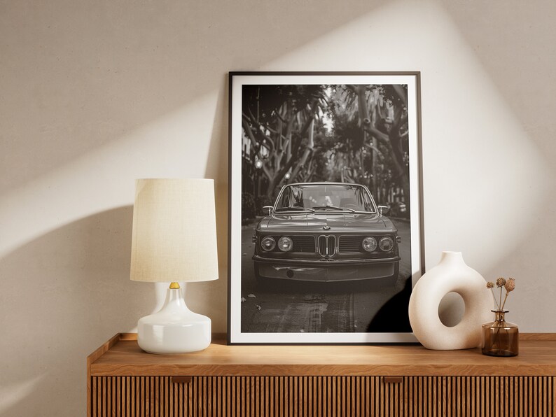 Puede incluir: Fotograf&iacute;a en blanco y negro de un coche cl&aacute;sico conduciendo por una calle bordeada de &aacute;rboles. El coche es un BMW vintage con una parrilla cromada y faros redondos. La foto est&aacute; enmarcada en un marco negro y est&aacute; colgada en una pared sobre una estanter&iacute;a de madera.