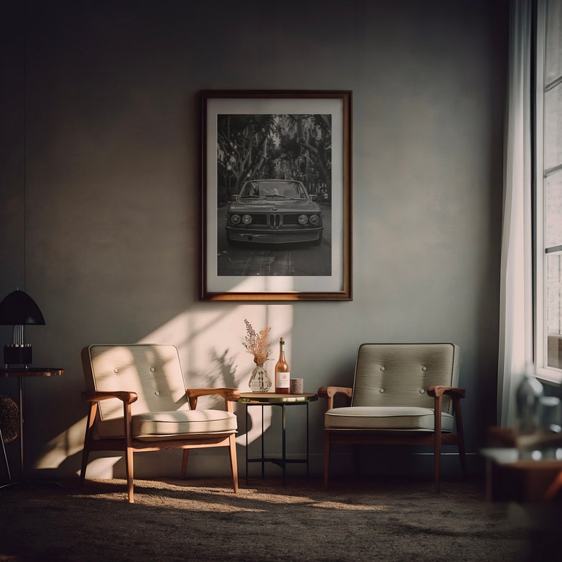 Puede incluir: Una fotograf&iacute;a en blanco y negro de un coche antiguo en una zona arbolada, enmarcada en un marco de madera marr&oacute;n y colgada en una pared gris. Dos sillones beige con marcos de madera est&aacute;n colocados frente a la foto enmarcada, uno frente al otro. Una peque&ntilde;a mesa de metal negro con una tapa de vidrio se encuentra entre las sillas, con un jarr&oacute;n de flores secas y dos botellas sobre la mesa.