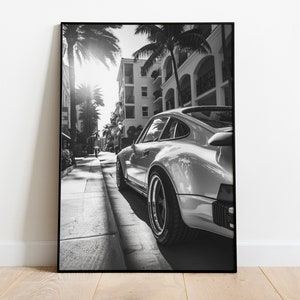 May include: A black and white photograph of a silver Porsche 911 driving down a street lined with palm trees and buildings.