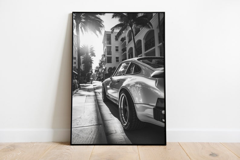 May include: A black and white photograph of a silver Porsche 911 driving down a street lined with palm trees and buildings.