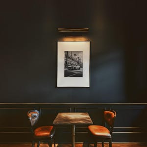 May include: A black and white photograph of a car driving down a street with palm trees. The photo is framed and hanging on a dark wall above a table with two chairs.