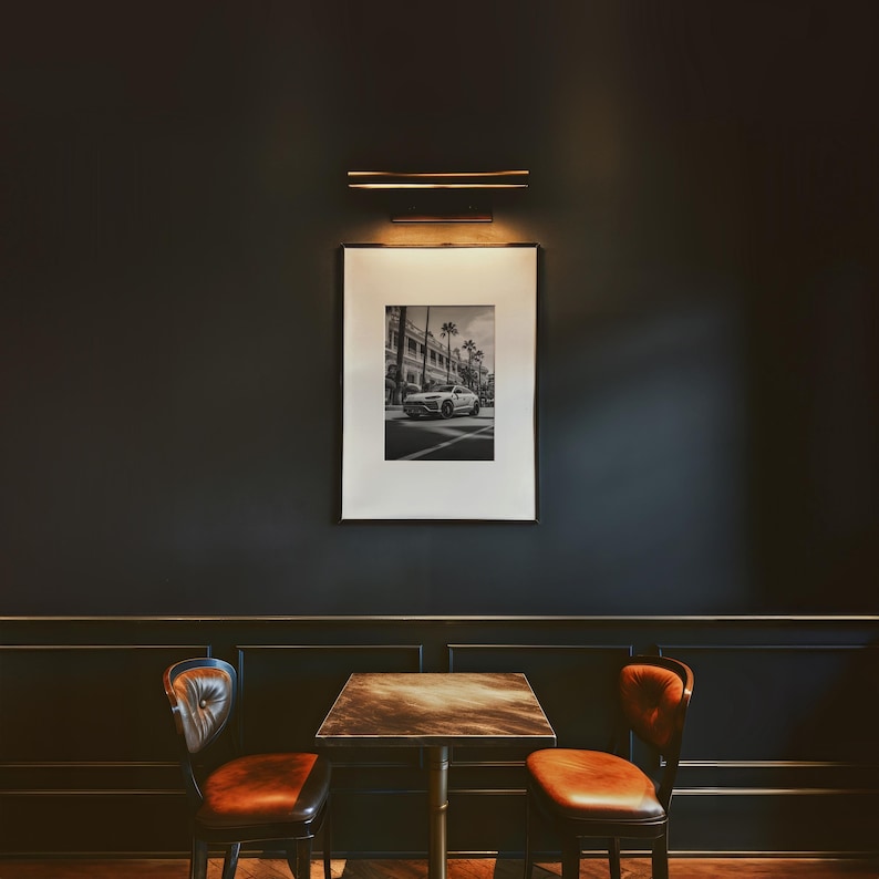 May include: A black and white photograph of a car driving down a street with palm trees. The photo is framed and hanging on a dark wall above a table with two chairs.