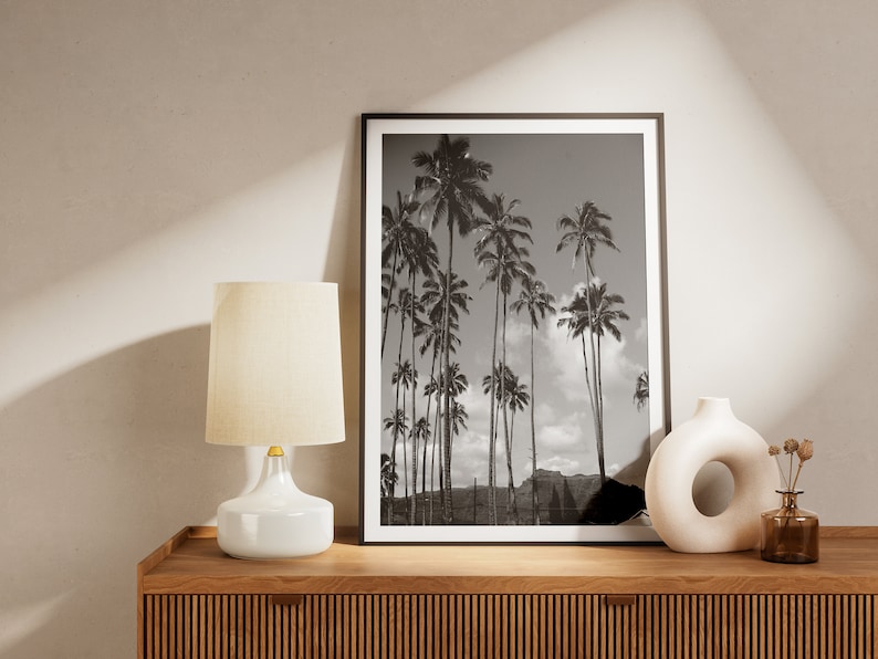 May include: A black and white photograph of palm trees against a cloudy sky is framed and displayed on a wooden shelf with a white lamp and a white ceramic vase with a hole in the side.