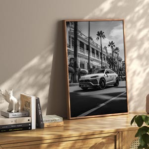 May include: Black and white photograph of a white Lamborghini SUV parked on a street in front of a building. The photo is framed in a wooden frame.