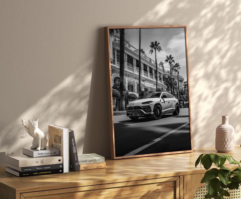 May include: Black and white photograph of a white Lamborghini SUV parked on a street in front of a building. The photo is framed in a wooden frame.