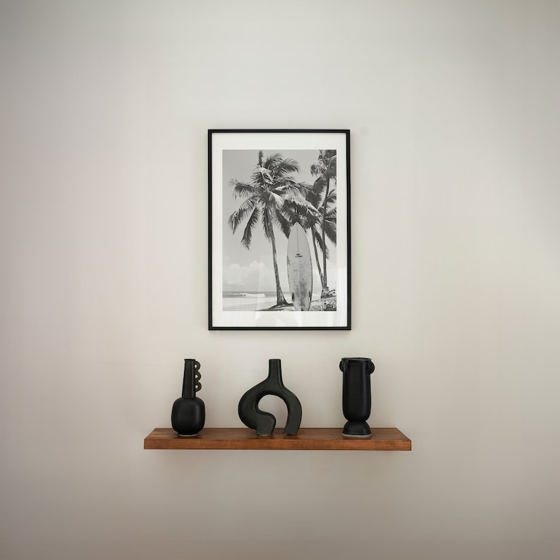May include: Black and white photograph of palm trees and a surfboard on a beach, framed in a black frame. Three black ceramic vases are displayed on a wooden shelf in front of the frame.