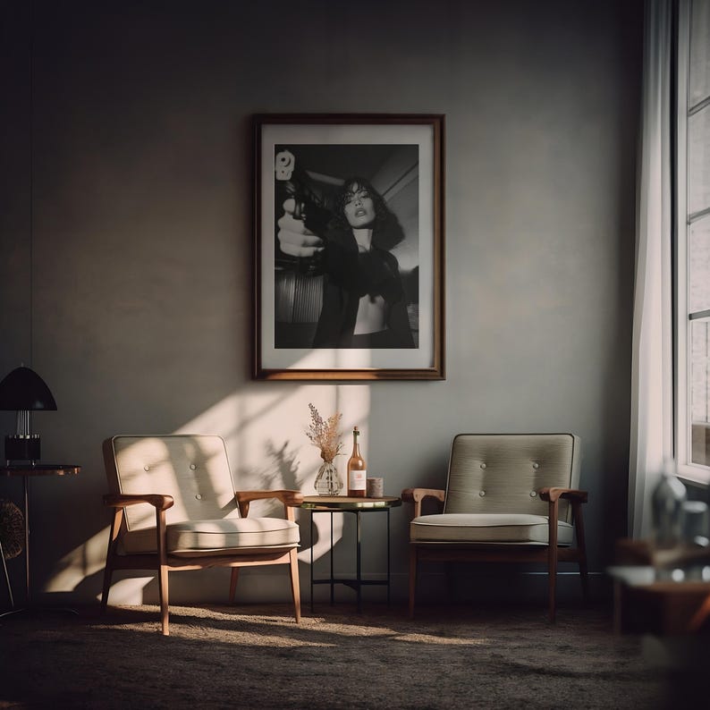 May include: A framed black and white photograph of a person holding a gun hangs on a gray wall. Two armchairs with light-colored cushions and wooden frames sit on a patterned rug. A small table holds a vase and a bottle.