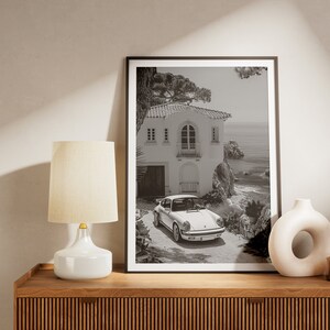 K&ouml;nnte beinhalten: Schwarzwei&szlig;foto eines wei&szlig;en Hauses mit Garagentor und einem davor geparkten Auto. Das Haus befindet sich auf einer Klippe mit Blick auf das Meer.