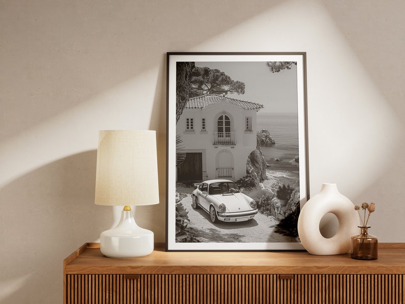 K&ouml;nnte beinhalten: Schwarzwei&szlig;foto eines wei&szlig;en Hauses mit Garagentor und einem davor geparkten Auto. Das Haus befindet sich auf einer Klippe mit Blick auf das Meer.