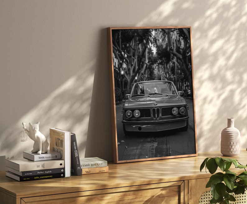 Puede incluir: Impresi&oacute;n fotogr&aacute;fica en blanco y negro de un coche cl&aacute;sico conduciendo por una calle bordeada de &aacute;rboles. El coche es un BMW vintage con una parrilla negra y faros redondos. La foto est&aacute; enmarcada en un marco de madera.