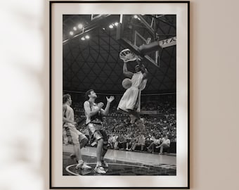 May include: Black and white photograph of a basketball game. A player in mid-air is about to dunk the ball. Another player is jumping to block the shot. The game is being played in a large arena with a crowd of spectators.