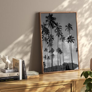 May include: Black and white photograph of palm trees against a cloudy sky. The photo is framed in a brown wood frame and is hanging on a wall.
