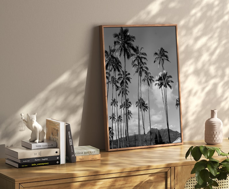 May include: Black and white photograph of palm trees against a cloudy sky. The photo is framed in a brown wood frame and is hanging on a wall.