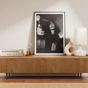 May include: A black and white framed print of a woman holding a gun, displayed on a wooden cabinet. The cabinet has a slatted design and is accompanied by a lamp, vases, and a stereo receiver. The room has a neutral color palette.