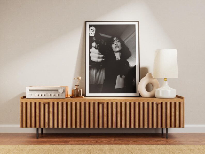 May include: A black and white framed print of a woman holding a gun, displayed on a wooden cabinet. The cabinet has a slatted design and is accompanied by a lamp, vases, and a stereo receiver. The room has a neutral color palette.
