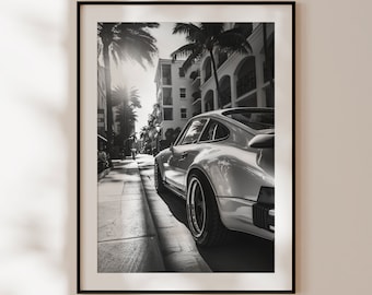 May include: A black and white photograph of a silver Porsche 911 driving down a street lined with palm trees and buildings.