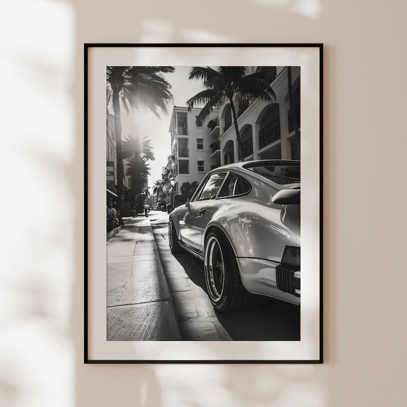 May include: A black and white photograph of a silver Porsche 911 driving down a street lined with palm trees and buildings.