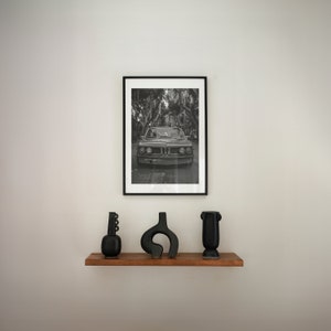 Puede incluir: Una fotograf&iacute;a en blanco y negro de un coche cl&aacute;sico conduciendo por una calle bordeada de &aacute;rboles. La foto est&aacute; enmarcada en un marco negro y est&aacute; colgada en una pared blanca. Tres jarrones de cer&aacute;mica negra est&aacute;n en un estante de madera debajo de la foto.