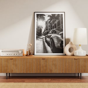 May include: A wooden media console with a black and white photo of a classic car in a city street above it. The console has a stereo receiver on top and a white table lamp with a white shade on the right side.