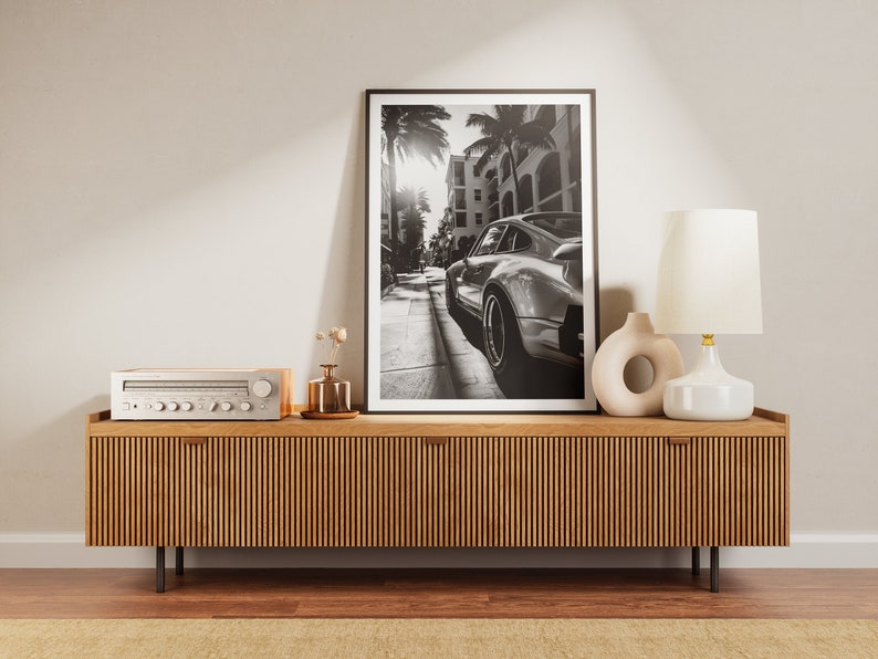 May include: A wooden media console with a black and white photo of a classic car in a city street above it. The console has a stereo receiver on top and a white table lamp with a white shade on the right side.