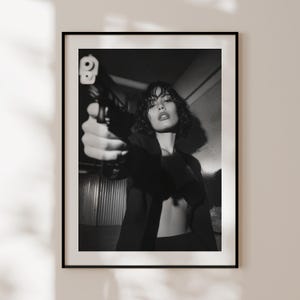 May include: Black and white framed print featuring a person holding a handgun, aiming directly at the viewer. The person is wearing a dark blazer and has short, curly hair. The image is a portrait with a dramatic, high-contrast aesthetic.
